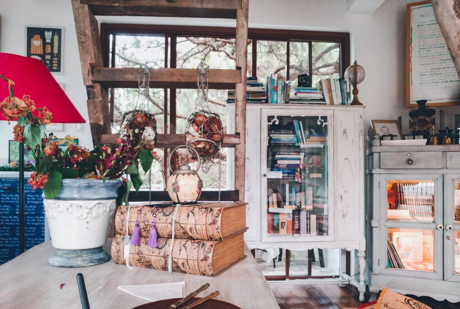 Estante envidraçada com livros antigos empilhados, um bule de porcelana e um vaso de flores em um canto de leitura doméstico